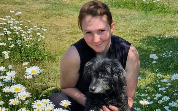 Mai Wölck, leidenschaftliche Tierosteopathin, kniet auf einer grünen Wiese, während ein Hund vor ihr steht. Eine Szene der Verbundenheit und Tiergesundheit.